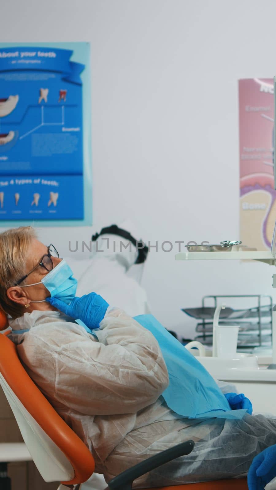 Senior patient talking with dentist doctor dressed in protective suit sitting in stomatological clinic during pandemic coronavirus. Nurse and orthodontist wearing coverall, face shield, mask gloves