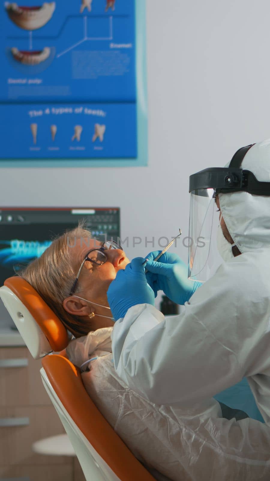 Senior woman sitting in dental clinic taking care of tooth health during covid-19 pandemic. Concept of new normal dentist visit in coronavirus outbreak wearing protective suit and face shield