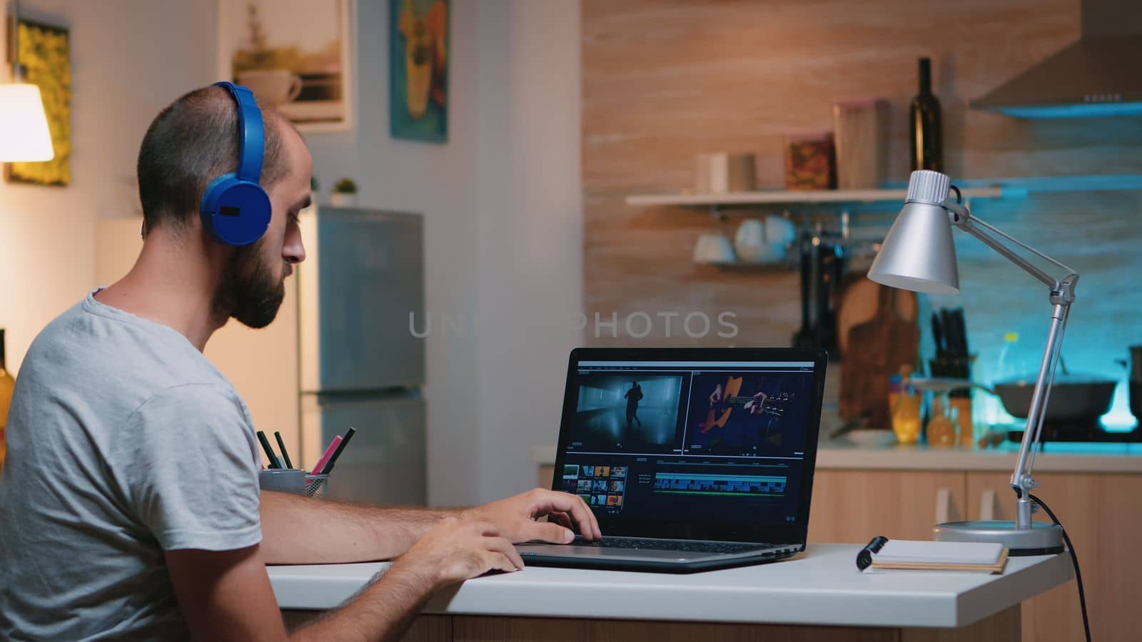 Professional videographer working in video editing app wearing headphones in front of laptop sitting in home kitchen. Freelancer processing audio film montage on professional laptop in midnight