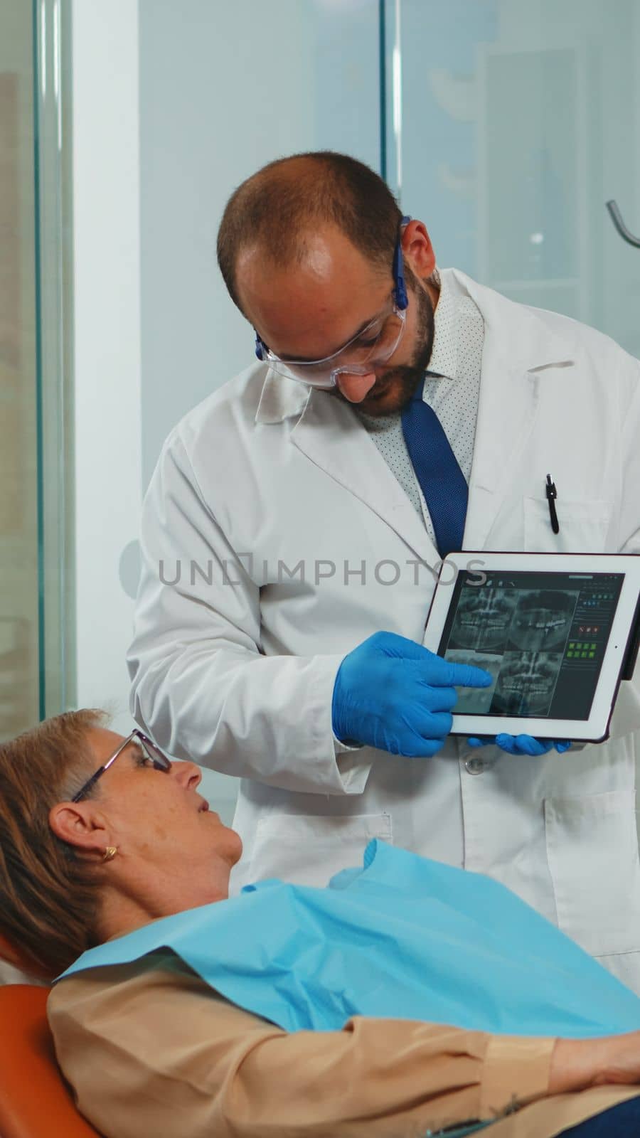 Orthodontist using tablet explaining dental x ray to patient sitting on dental chair in stomatologic office. Dentist showing to old woman radiography using digital device working in modern clinic.