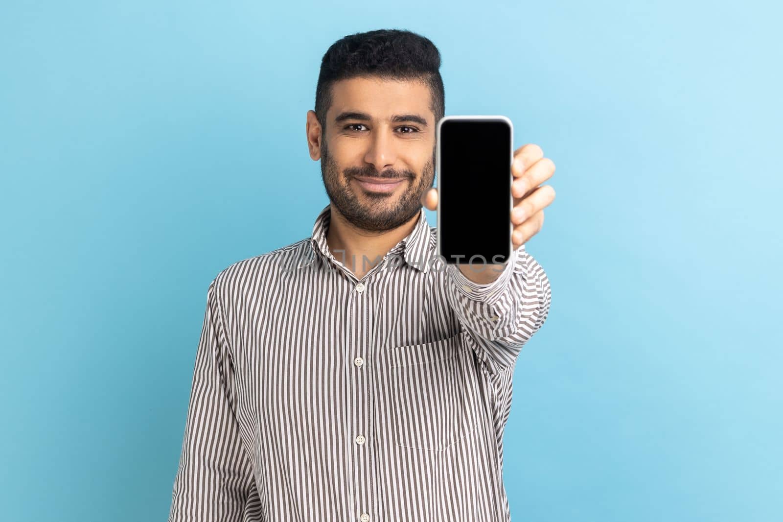 Portrait of bearded businessman holding out cellphone and smiling at camera, recommending gadget or mobile application, wearing striped shirt. Indoor studio shot isolated on blue background.