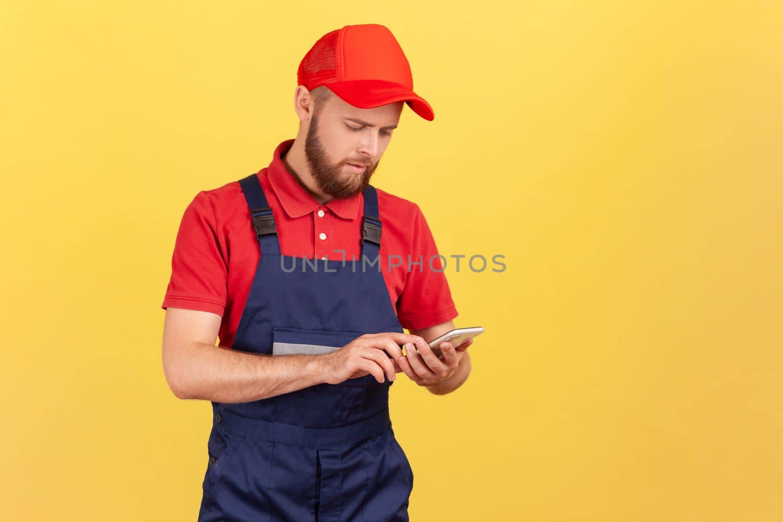 Serious handyman in overalls typing on mobile phone, using cellphone messenger to accept online order as delivery, repair and maintenance services. Indoor studio shot isolated on yellow background.