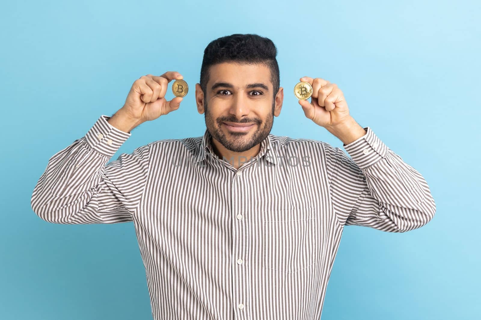 Cryptocurrency btc coin. Rich satisfied happy businessman looking at camera, holds golden bitcoin, electronic money, wearing striped shirt. Indoor studio shot isolated on blue background.