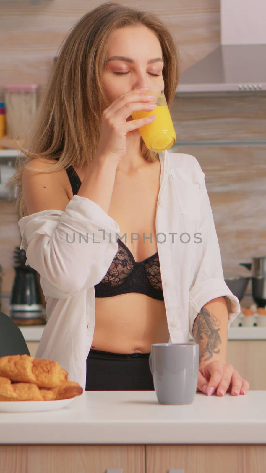 Seductive woman in sexy underwear enjoing the morning drinking a glass of fresh orange juice sitting in the kitchen. Young blode lady with tattoos wearing black lingerie refreshing sunday morning
