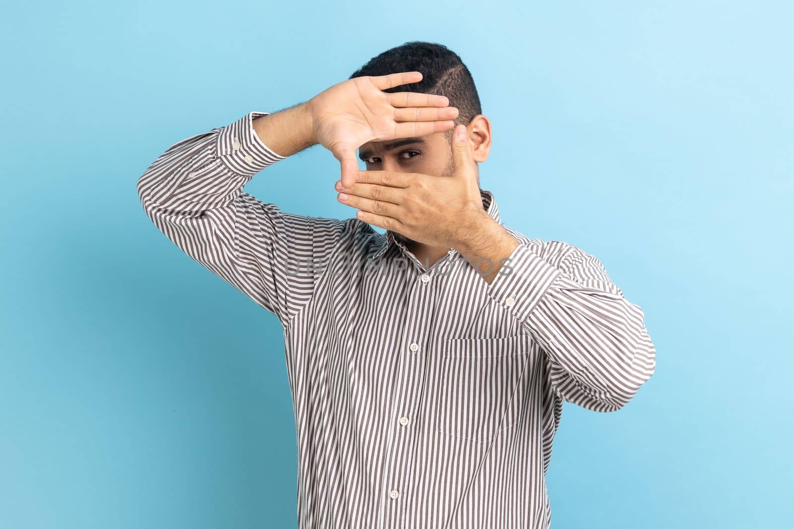 Creative businessman looking through hand frame gesture, cropping image, zooming and focusing on distance, viewing target, wearing striped shirt. Indoor studio shot isolated on blue background.