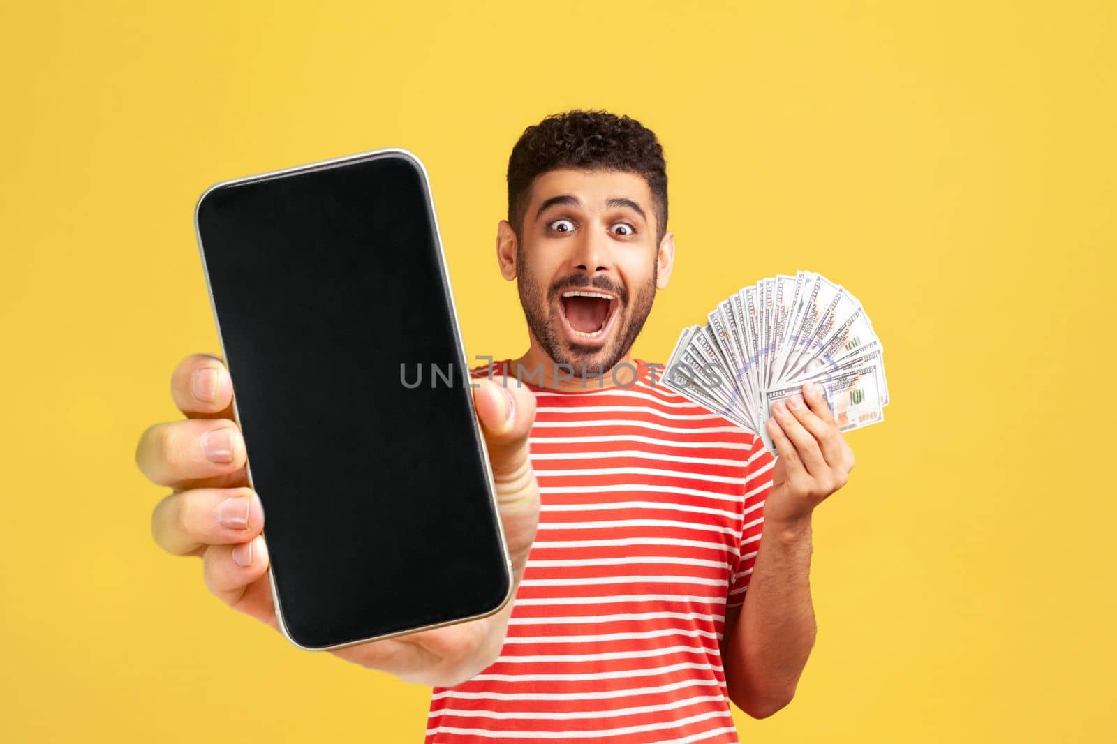 Surprised happy man with beard in red T-shirt, looking with big amazed eyes and open mouth, holding money and showing mobile phone with empty screen. Indoor studio shot isolated on yellow background.