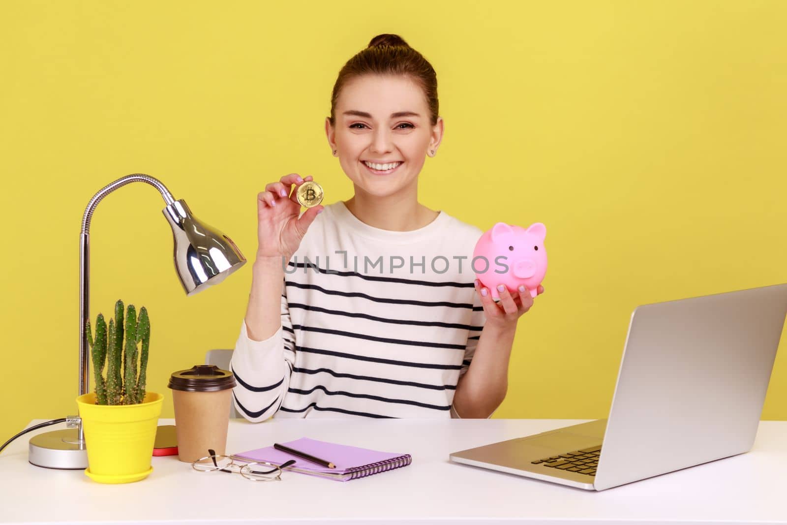 Portrait of satisfied dark haired woman investing in bitcoins, putting golden crypto coin into piggy bank and looking at camera. Indoor studio studio shot isolated on yellow background.
