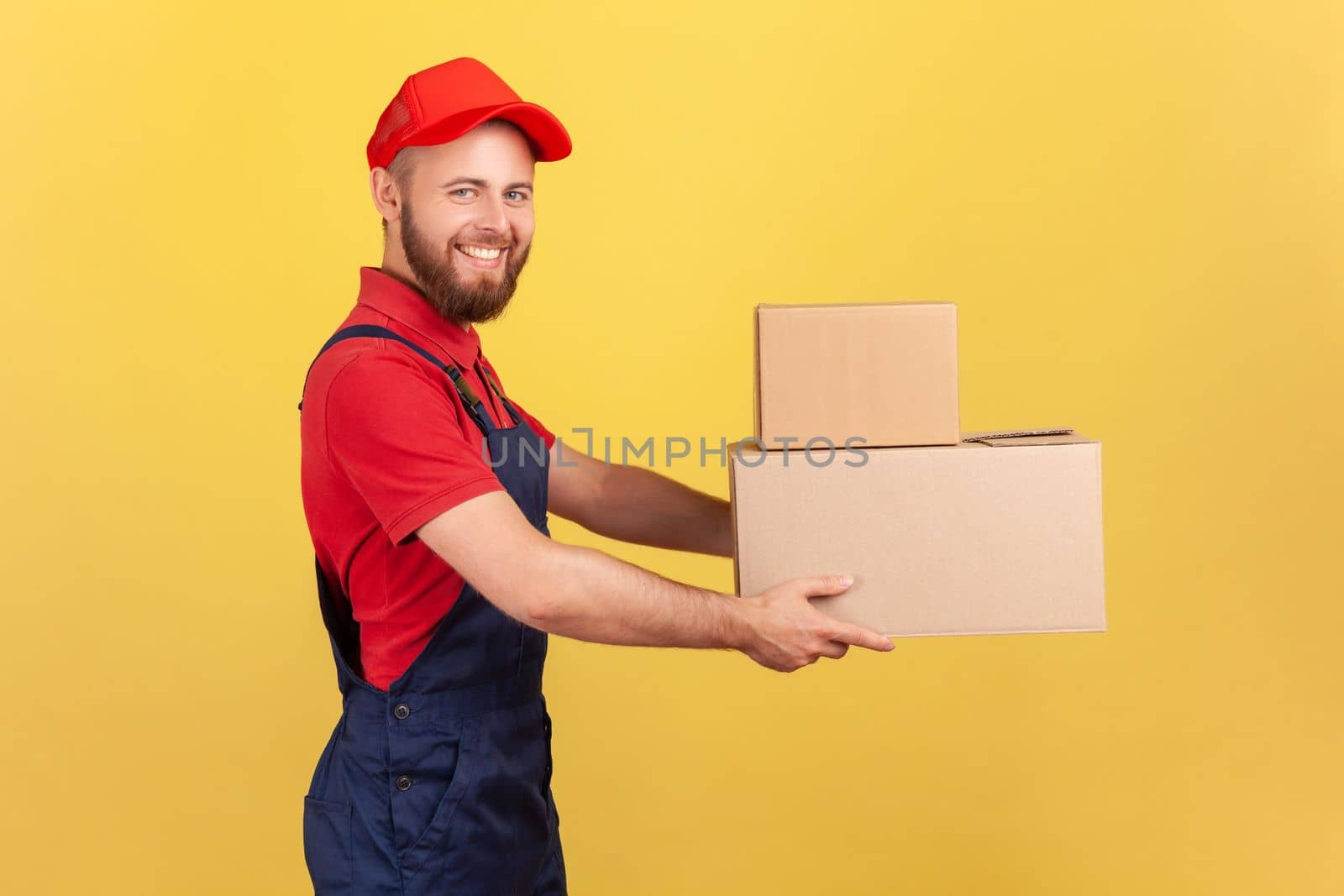 Side view of happy deliveryman holding two cardboard boxes, delivering order door-to-door, shipment and cargo transportation service. Indoor studio shot isolated on yellow background.