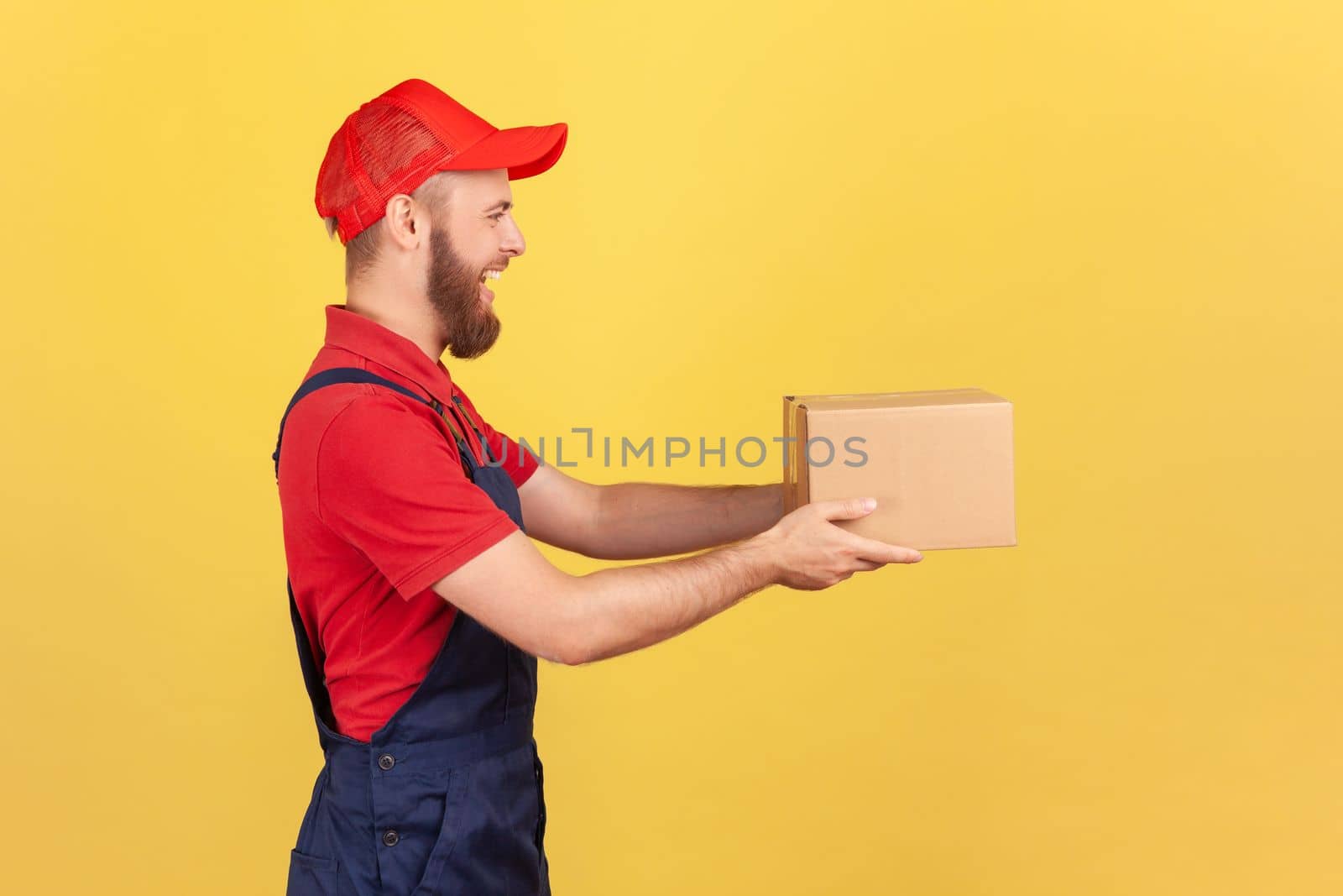 Profile portrait of positive deliveryman holding cardboard parcel, delivering order door-to-door, shipment and cargo transportation service. Indoor studio shot isolated on yellow background.