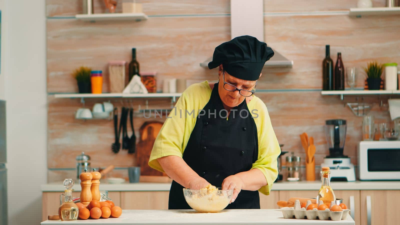 Woman wearing chef bonete while mixing cracked eggs with flour in kitchen while preparing food following traditional recipe. Retired elderly baker kneading in glass bowl ingredients for homemade cake
