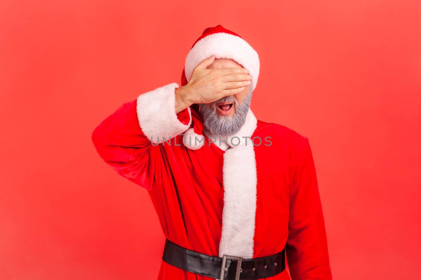 Portrait of elderly man with gray beard wearing santa claus costume covering eyes with palm, does not want to see, avoiding problems. Indoor studio shot isolated on red background.