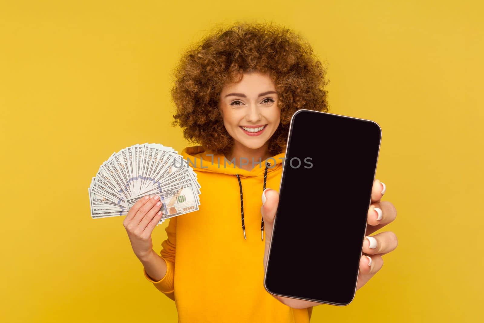Portrait of satisfied smiling woman with Afro hairstyle wearing hoodie holding fan of dollar banknotes and showing big smart phone empty display. Indoor studio shot isolated on yellow background.