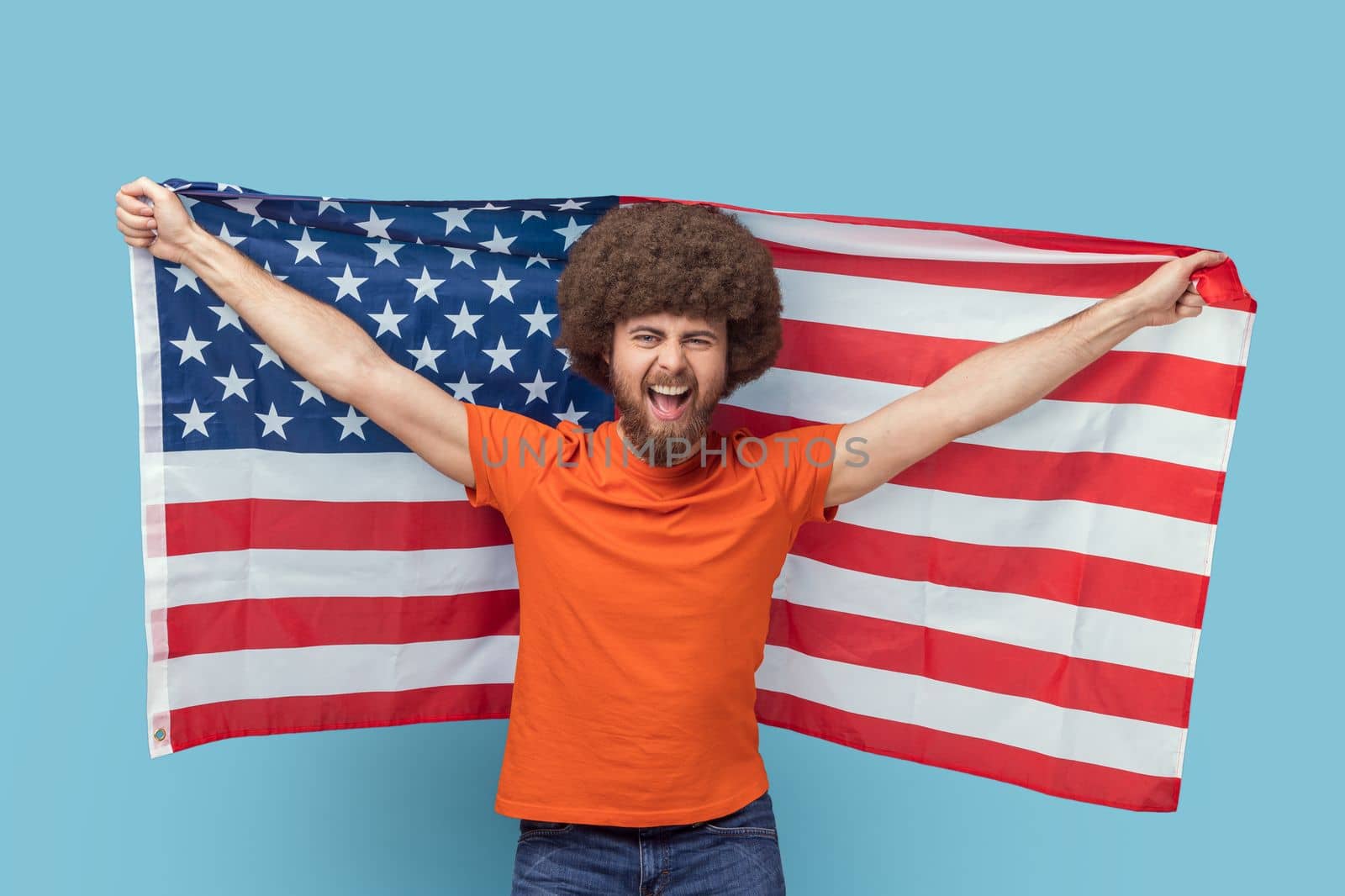 Portrait of man with Afro hairstyle in T-shirt holding USA flag and looking at camera with rejoicing look, screaming, celebrating national holiday. Indoor studio shot isolated on blue background.