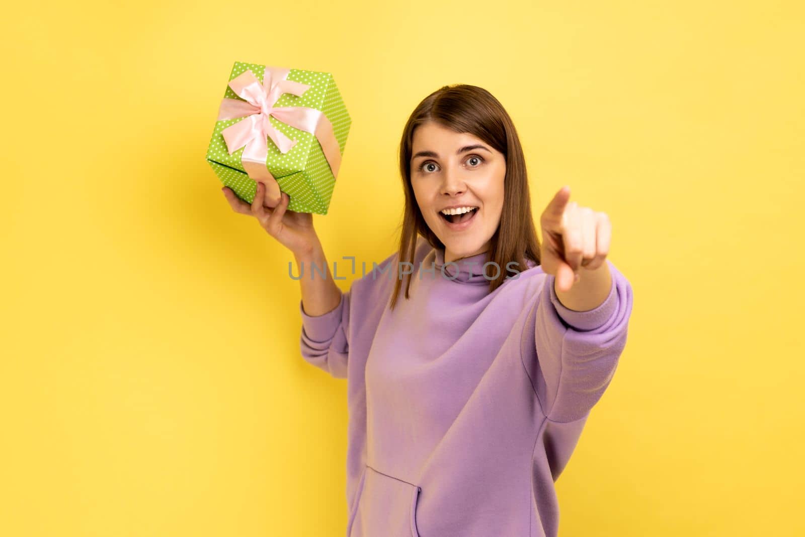 Happy excited woman throwing present box, having happy look, celebrating holiday, congratulating and pointing to camera, wearing purple hoodie. Indoor studio shot isolated on yellow background.