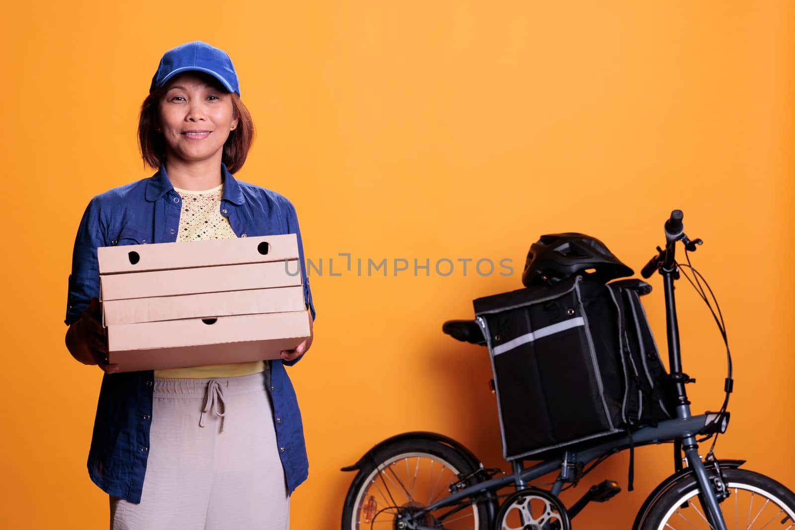 Restaurant worker wearing blue uniform holding stack of pizza delivering to customer on bike. Food delivery employee standing in studio with yellow background. Food service and transportation