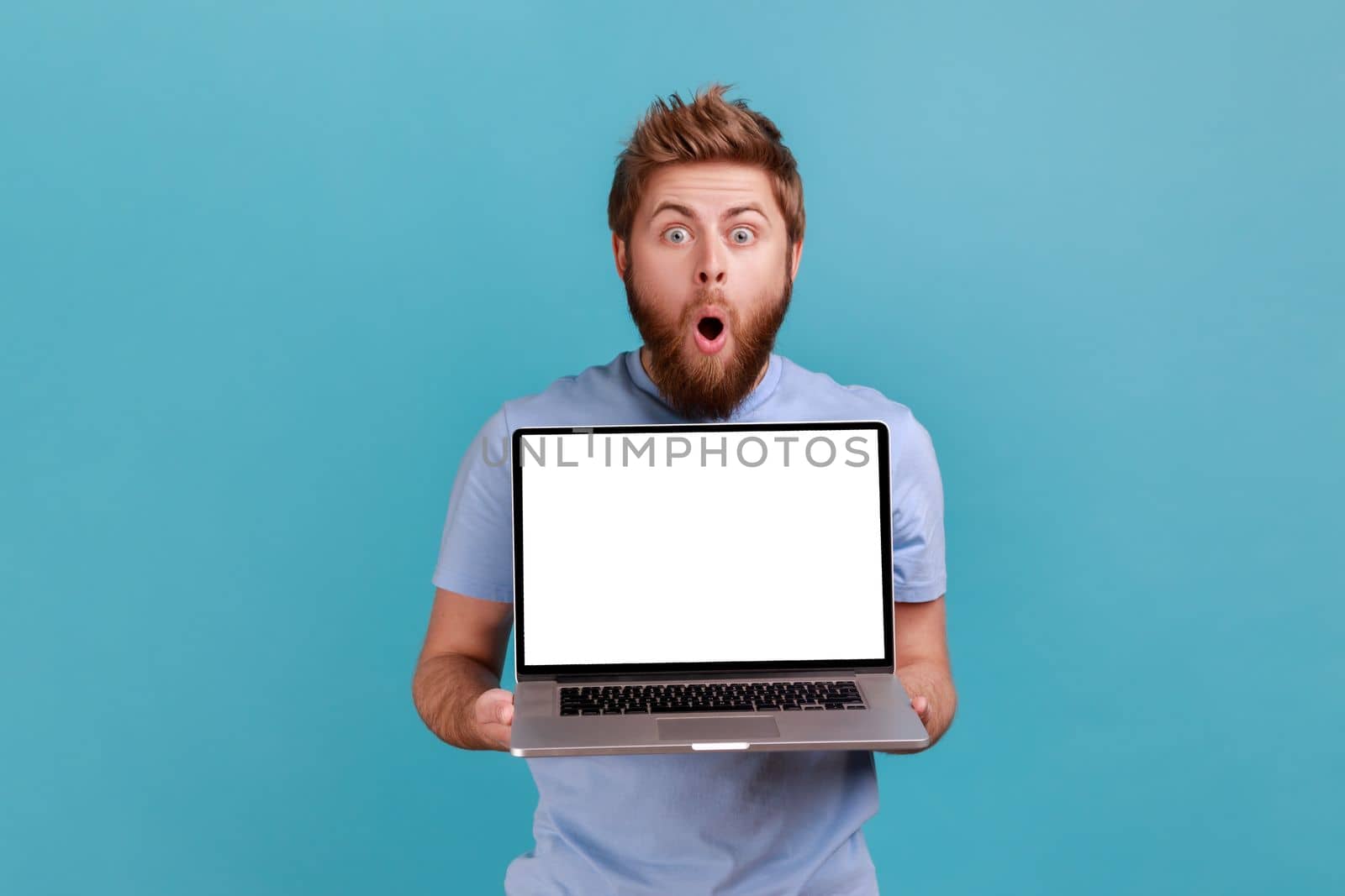 Portrait of bearded man holding and showing laptop with blank empty display, place for your advertisement, looking at camera with big eyes and open mouth Indoor studio shot isolated on blue background