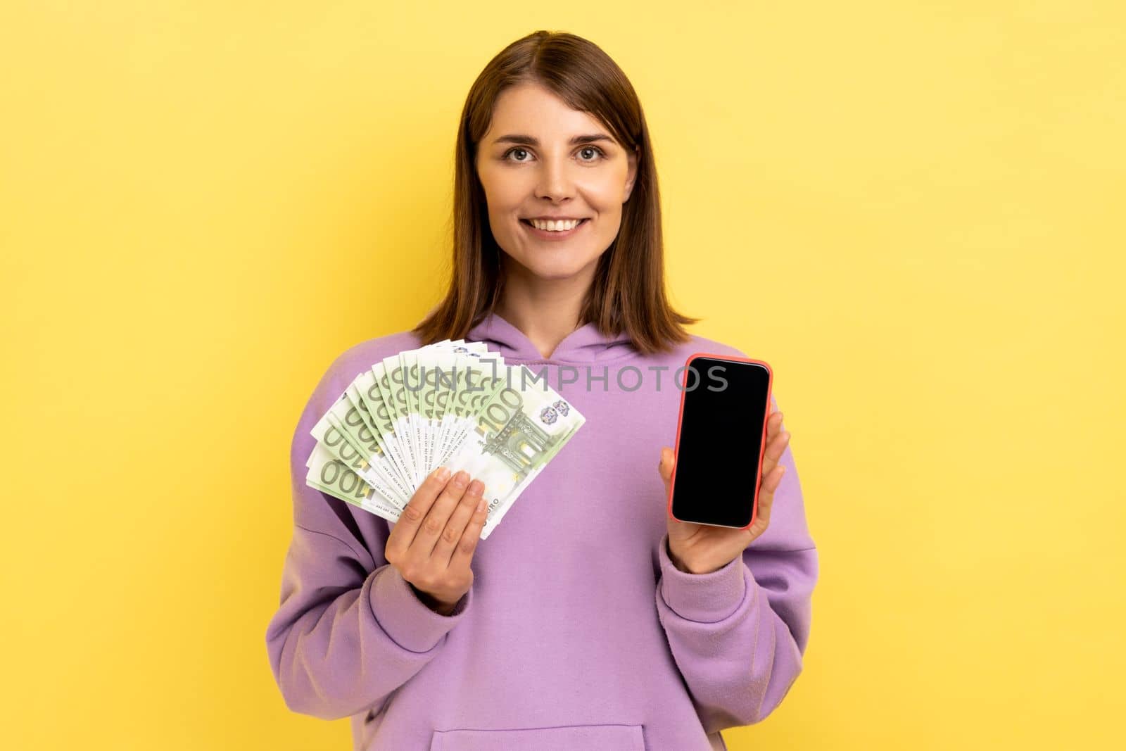 Positive satisfied dark haired woman holding euro banknotes and cell phone with empty screen, online betting, winning, wearing purple hoodie. Indoor studio shot isolated on yellow background.
