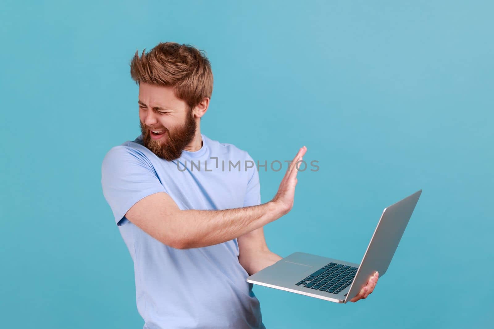 Portrait of handsome bearded man showing stop gesture, avoiding watch something unpleasant, turning face from display, forbidden content. Indoor studio shot isolated on blue background.