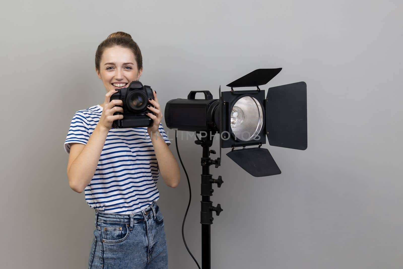 Portrait of smiling satisfied woman photographer wearing striped T-shirt standing with photocamera and reflector, looking at camera. Indoor studio shot isolated on gray background.