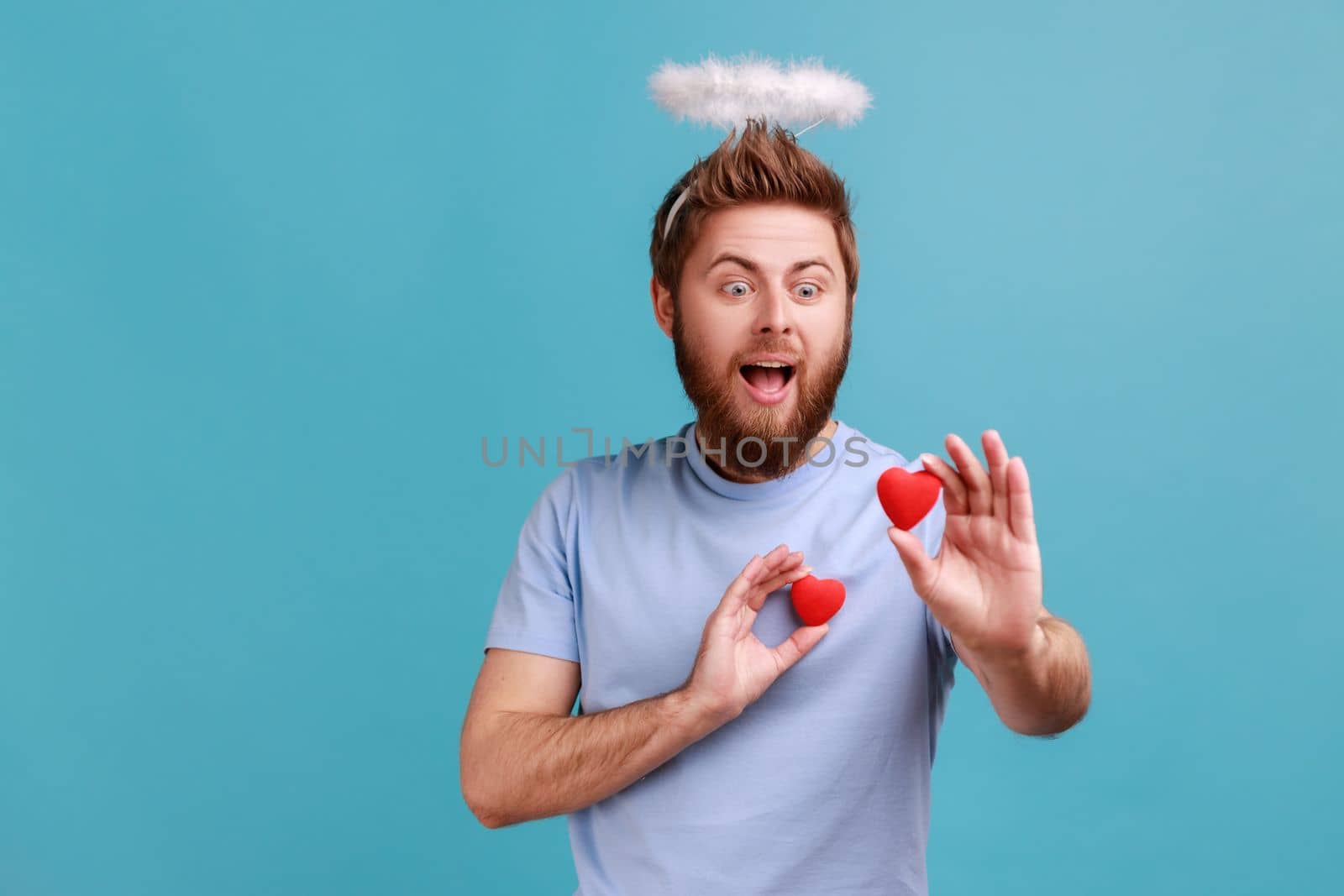 Portrait of excited bearded angelic man with holy nimbus on his head holding two red toy hears, expressing love and romantic feelings. Indoor studio shot isolated on blue background.