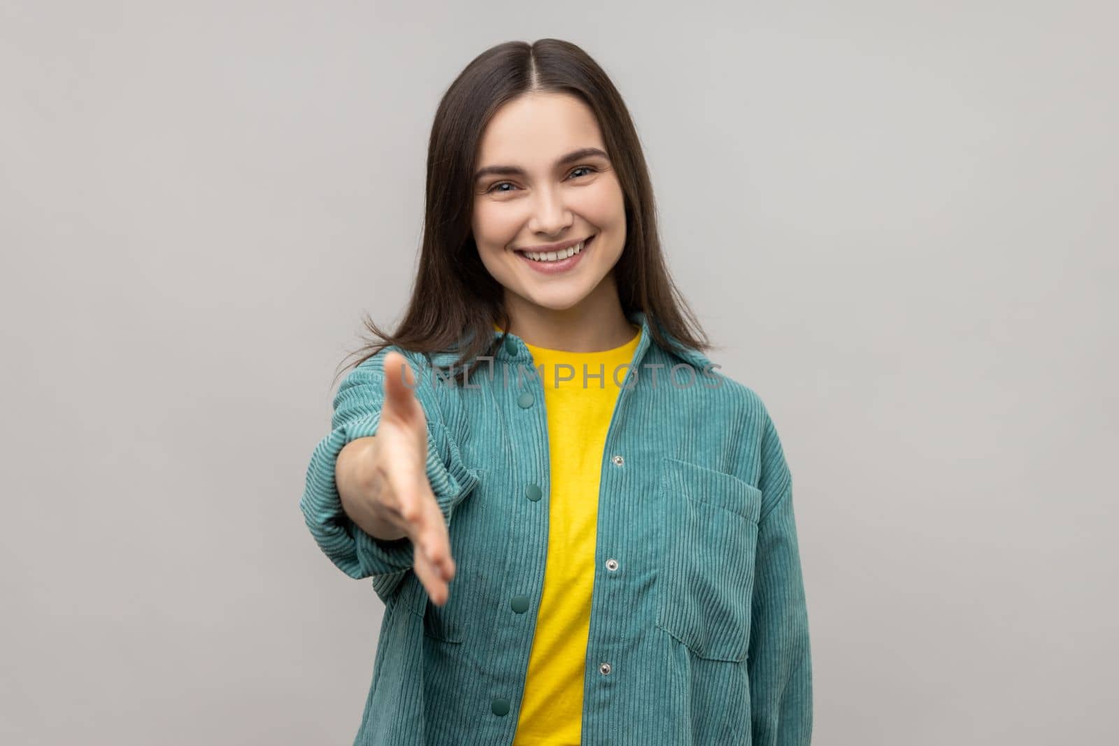 Portrait of happy beautiful young adult woman standing and giving hand to greeting and toothy smile, wearing casual style jacket. Indoor studio shot isolated on gray background.