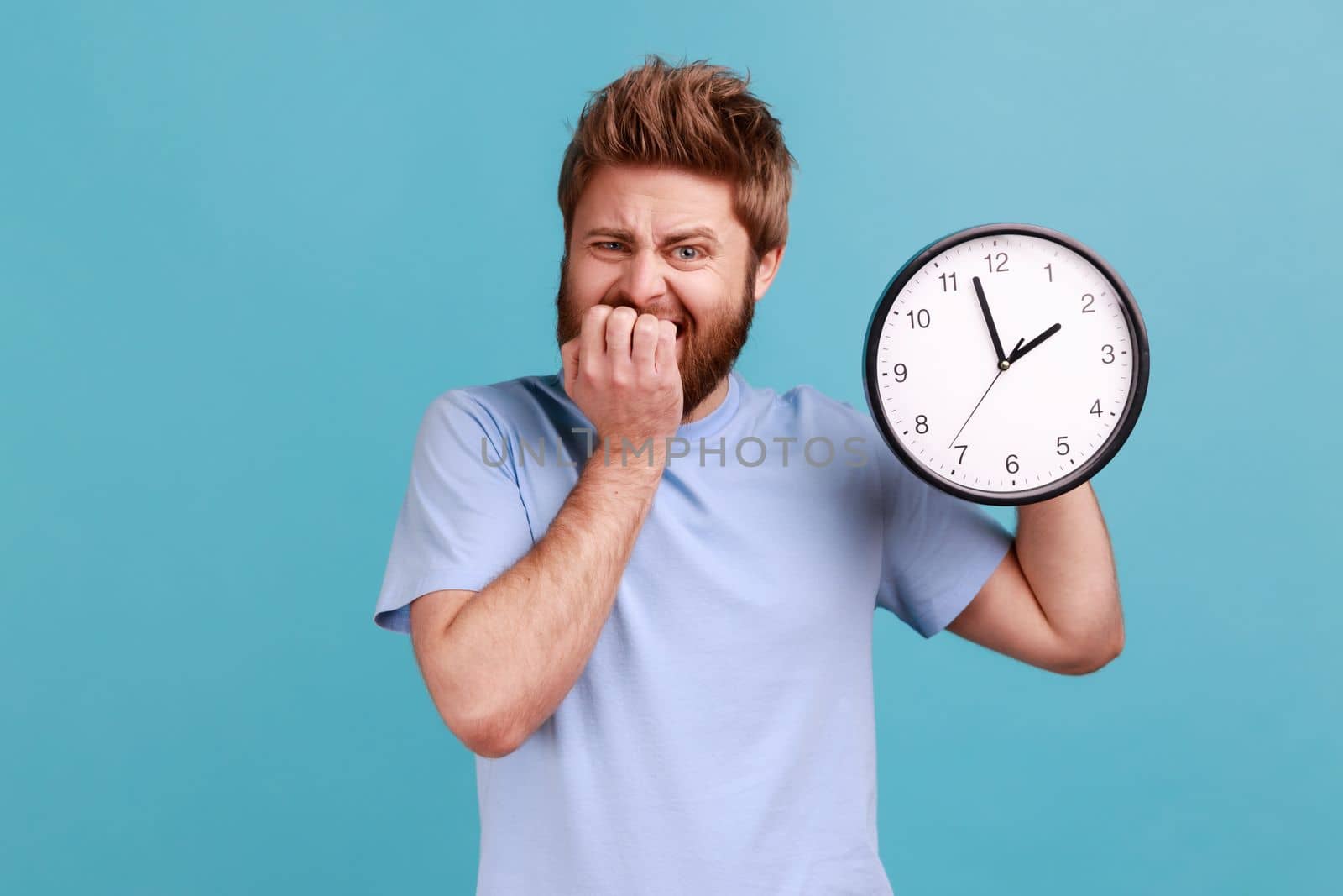 Portrait of nervous depressed confused young adult bearded man biting nails holding big wall clock, deadline, need hurry up, looking at camera. Indoor studio shot isolated on blue background.