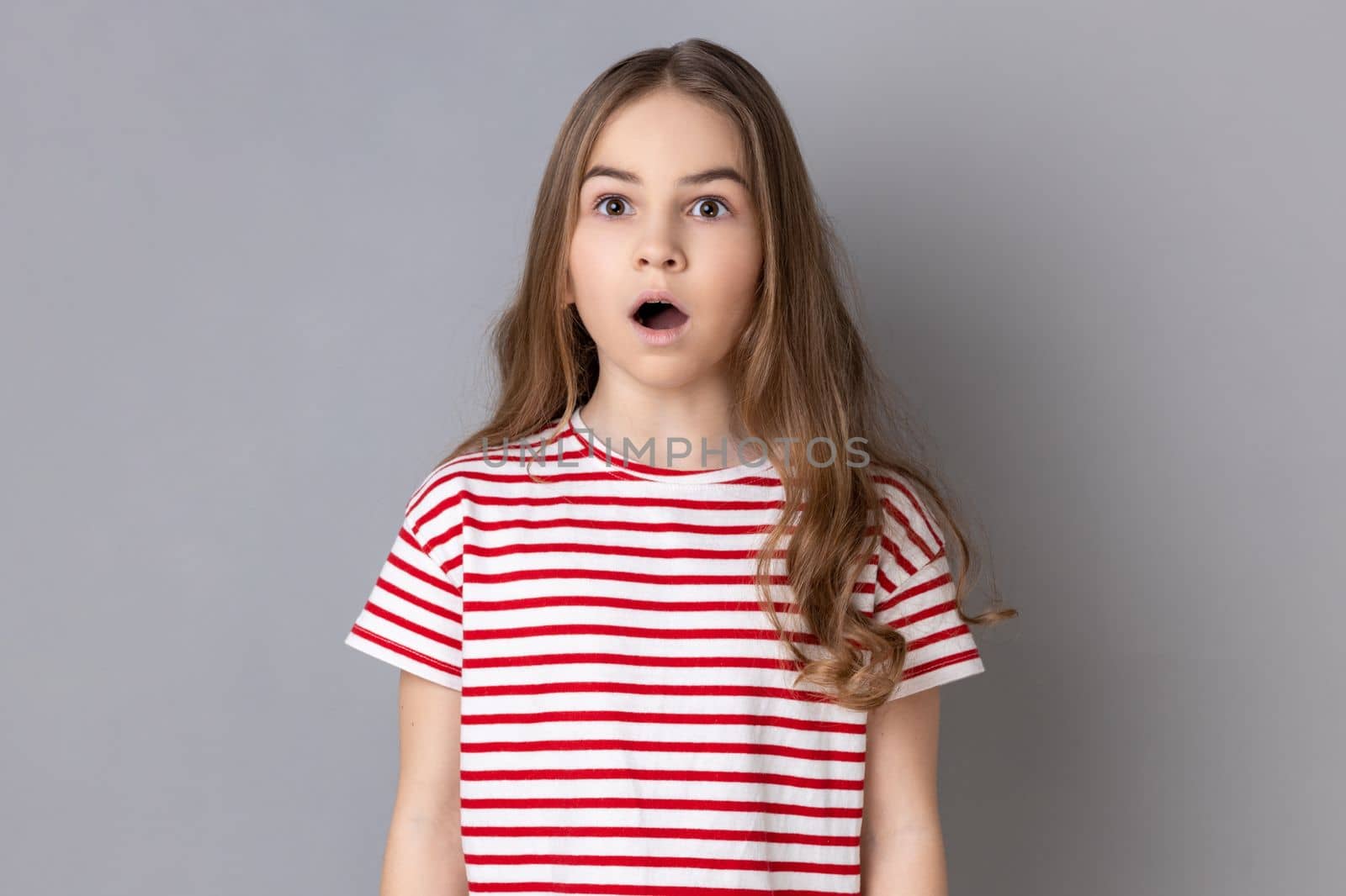 Portrait of astonished surprised little girl wearing striped T-shirt looking with shocked expression at camera, keeping mouth widely open. Indoor studio shot isolated on gray background.