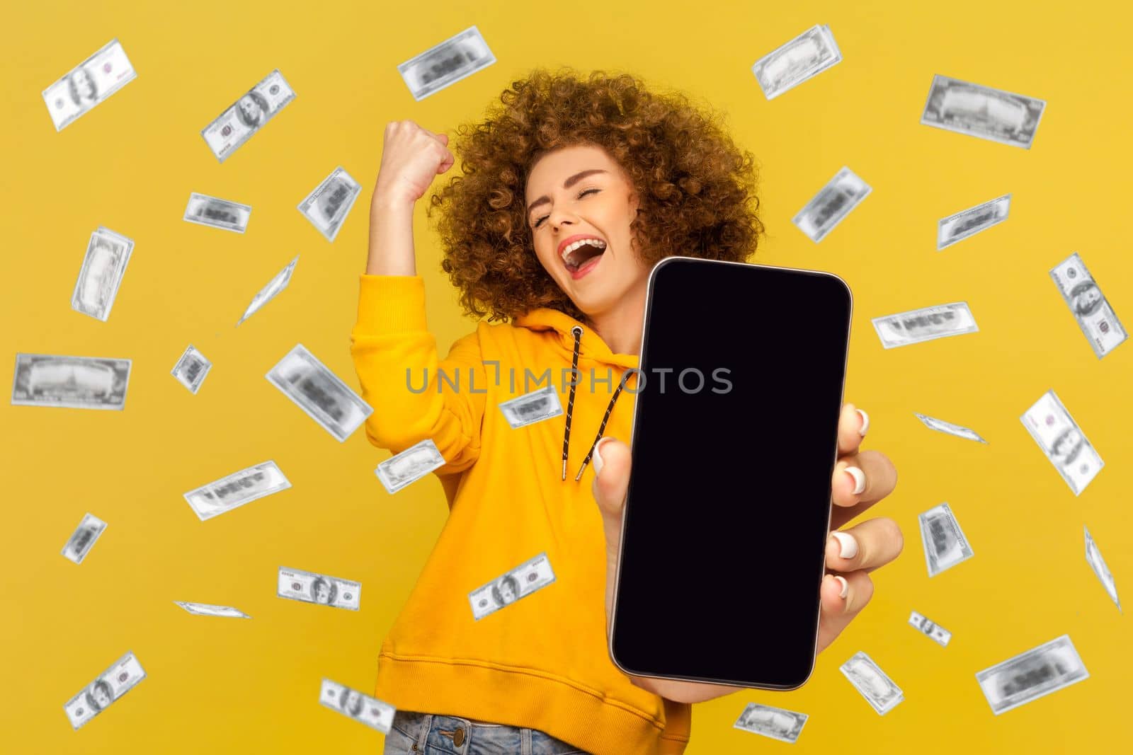 Excited woman with Afro hairstyle showing big empty screen for promotional text or advertisement, rejoicing her winning, standing in money rain. Indoor studio shot isolated on yellow background.