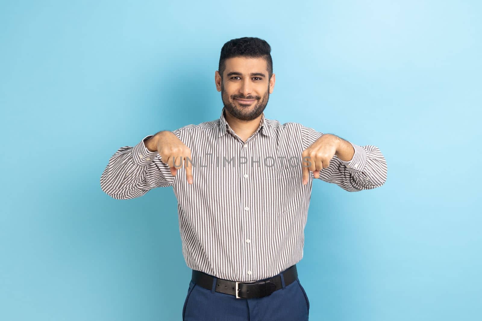 Cheerful satisfied businessman pointing fingers down, paying attention to advertising area, blogger recommending to subscribe, wearing striped shirt. Indoor studio shot isolated on blue background.