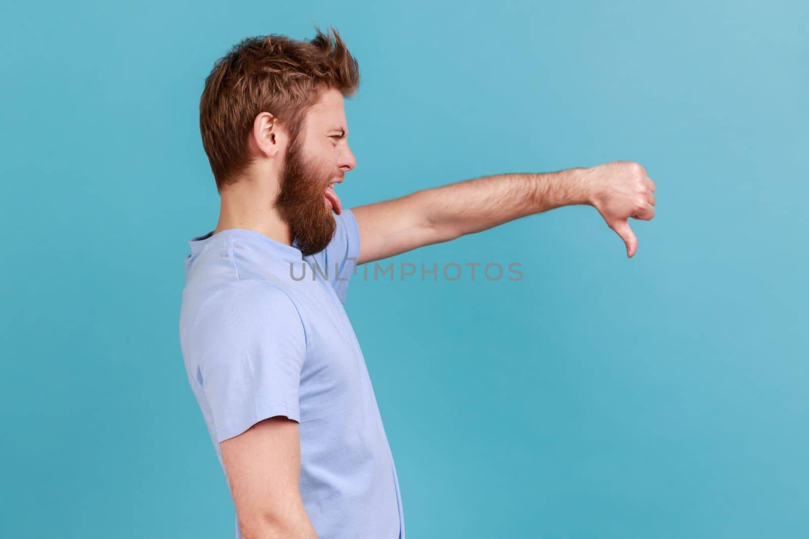 Side view of bearded man criticizing bad quality with thumbs down displeased grimace, showing dislike gesture, expressing disapproval. Indoor studio shot isolated on blue background.
