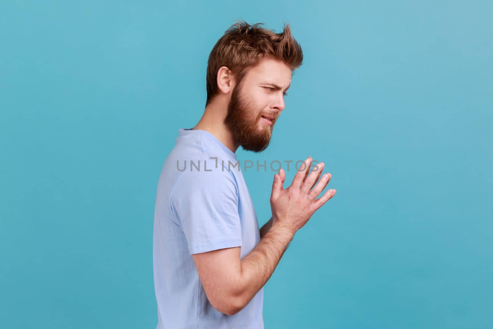 Side view of cunning bearded man schemes something keeps hands together and makes plans, sly expression looking at camera thoughtfully. Indoor studio shot isolated on blue background.