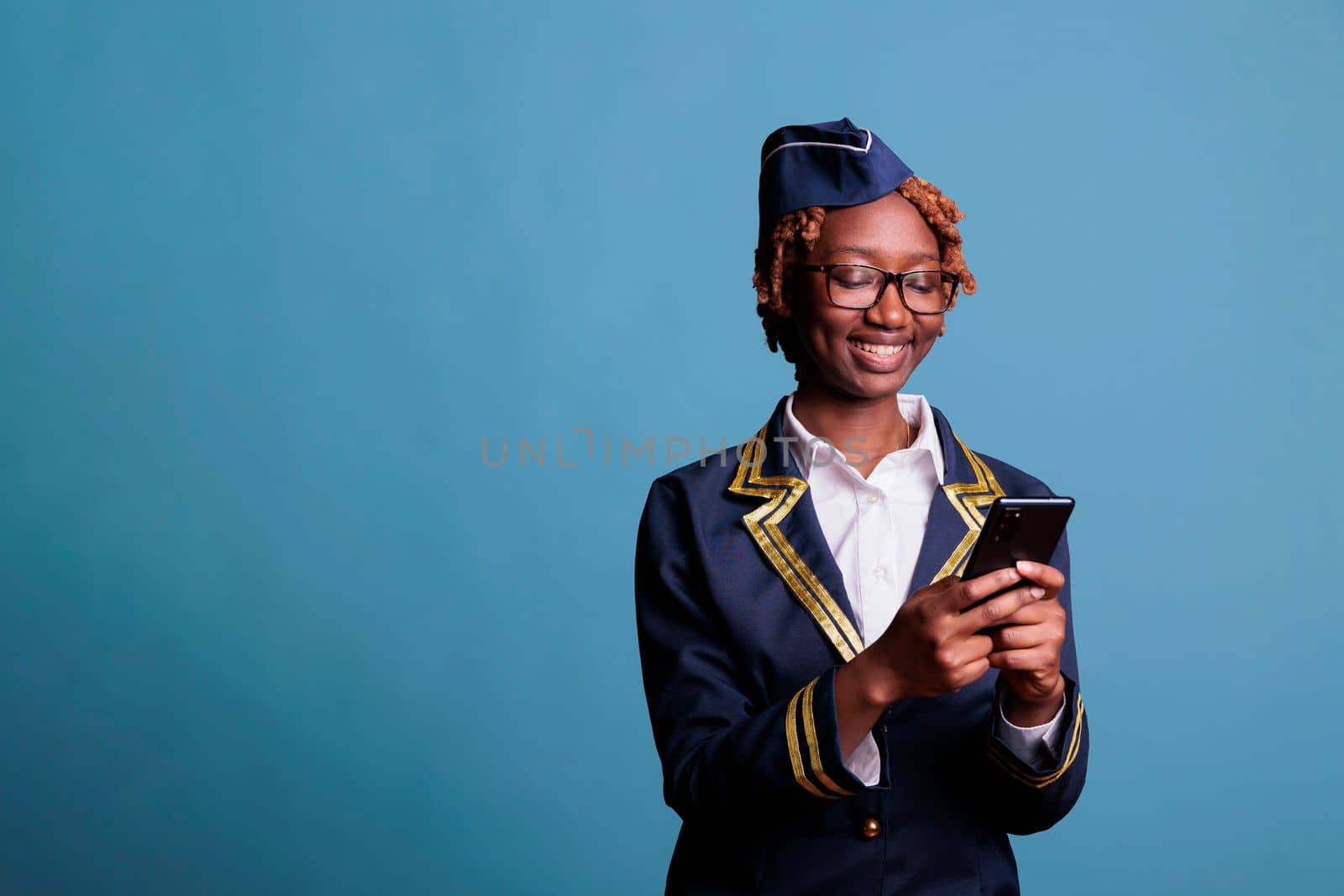 Smiling female flight attendant wearing uniform reading messages on mobile phone. Entertained happy stewardess with handheld device in studio shot against blue background.