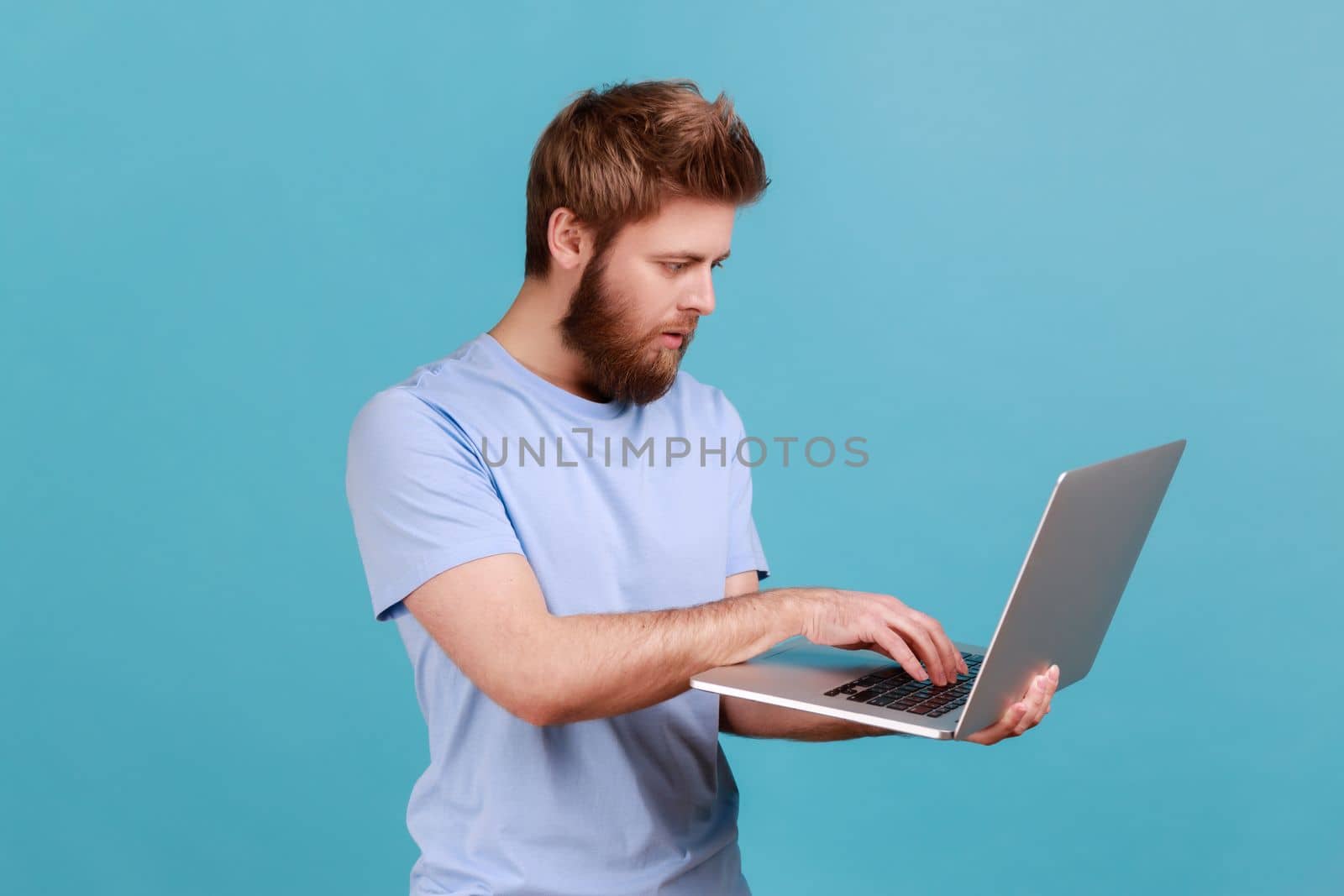 Portrait of bearded man holding wireless computer in hand, working on laptop, making edits, deadline, having serious facial expression. Indoor studio shot isolated on blue background.
