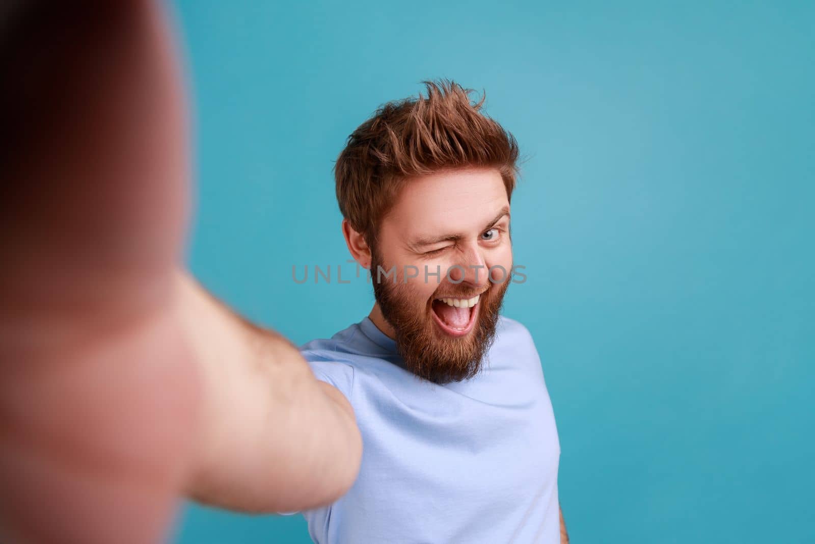 Portrait of excited optimistic bearded man taking selfie POV, winking to camera, being in excellent mood, flirting, point of view photo. Indoor studio shot isolated on blue background.
