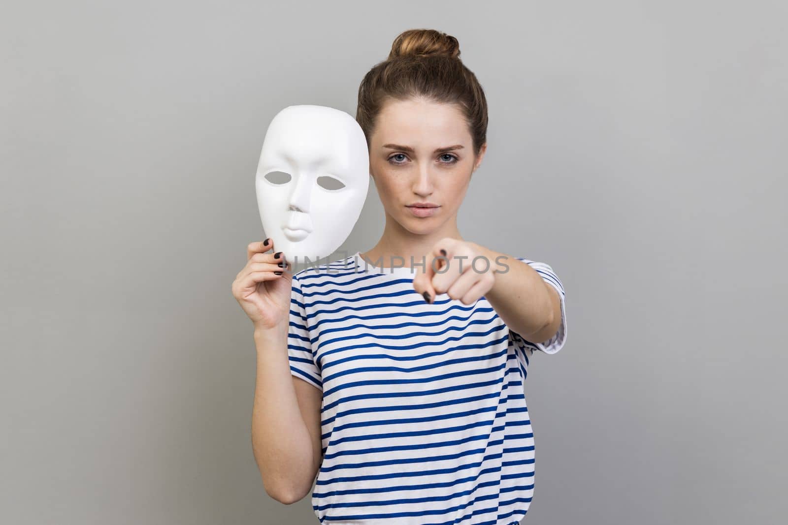 Portrait of serious strict woman wearing striped T-shirt holding white mask in hand, pointing finger to camera with serious expression. Indoor studio shot isolated on gray background.