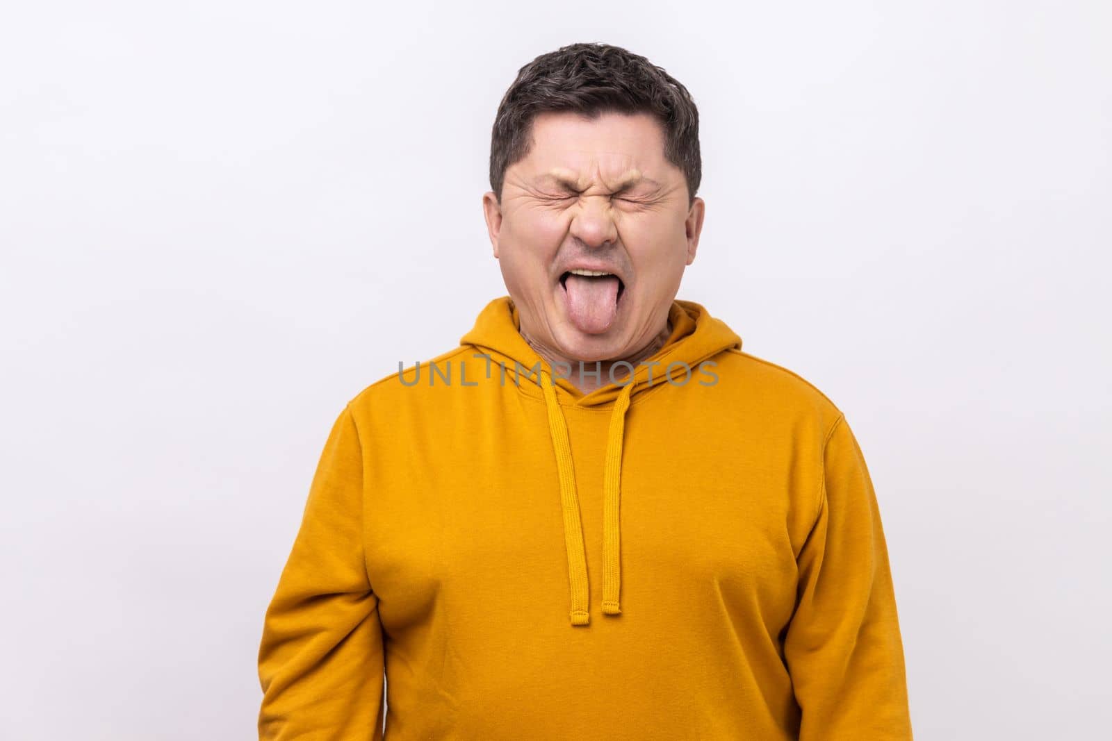 Naughty infantile middle aged man closing eyes and showing tongue, having fun, fooling around, childish manners, wearing urban style hoodie. Indoor studio shot isolated on white background.