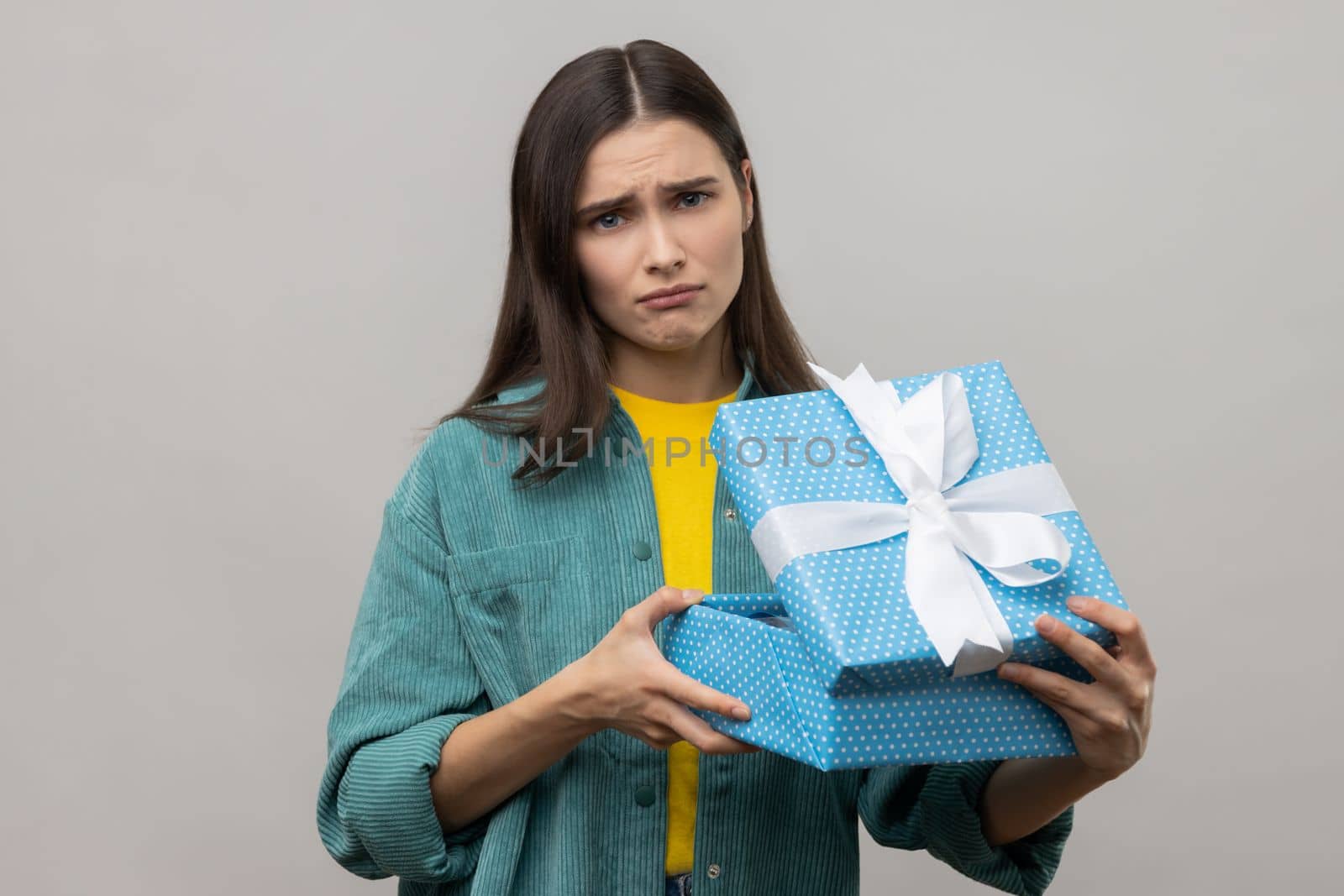 Sad upset woman with dark hair unboxing blue gift box, looking at camera with sorrow facial expression, birthday present, wearing casual style jacket. Indoor studio shot isolated on gray background.