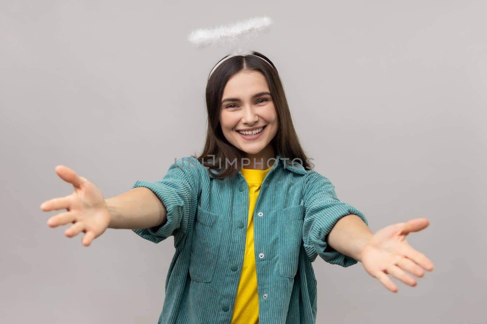 Come into my hands. Attractive angelic woman with halo over head stretching arms to camera with kind friendly smile, going to embrace. Indoor studio shot isolated on gray background.