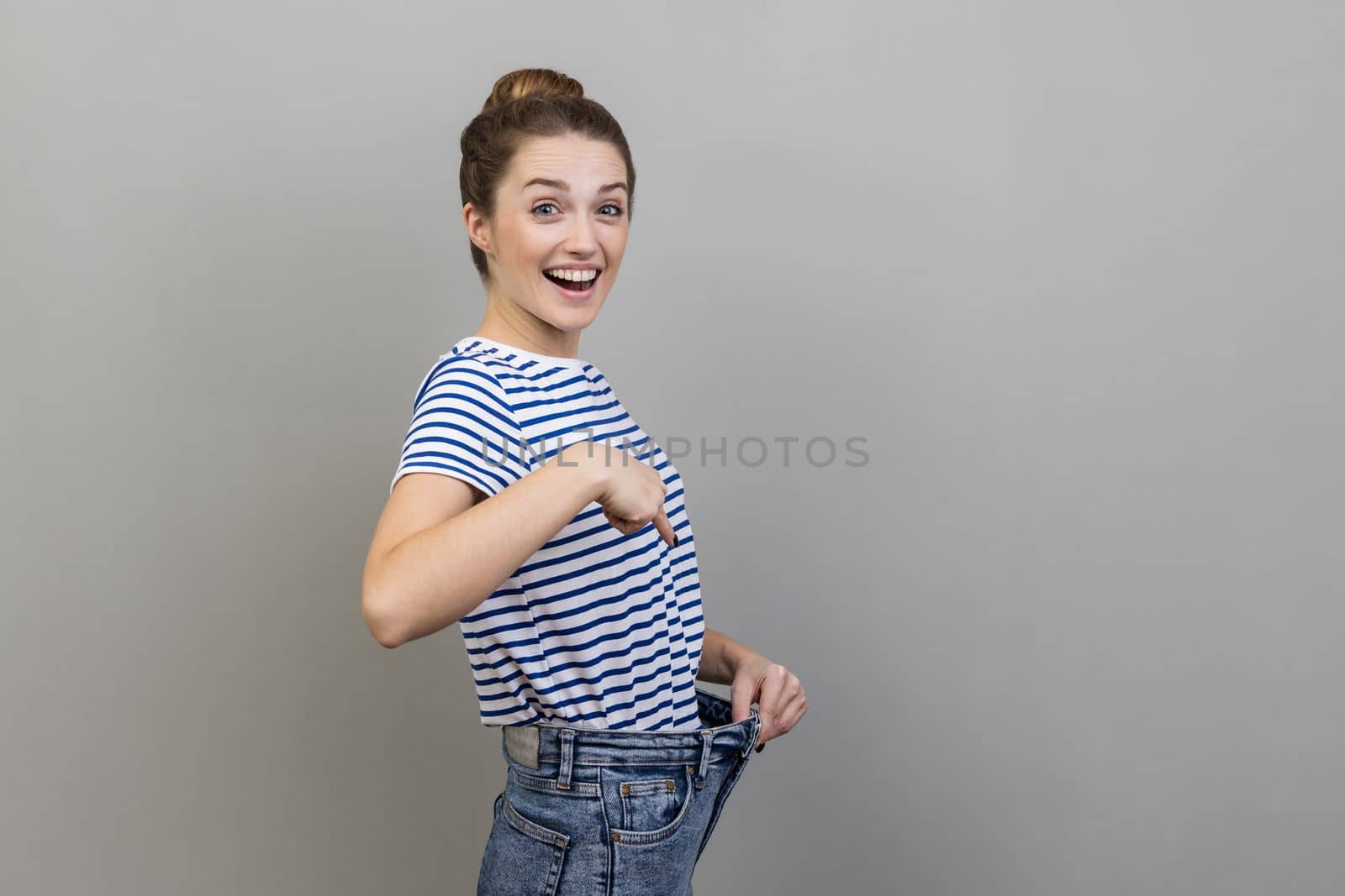 Attractive woman wearing striped T-shirt have willpower to loose weight, pointing at slim waist and pointing finger on big trousers, diet conception. Indoor studio shot isolated on gray background.