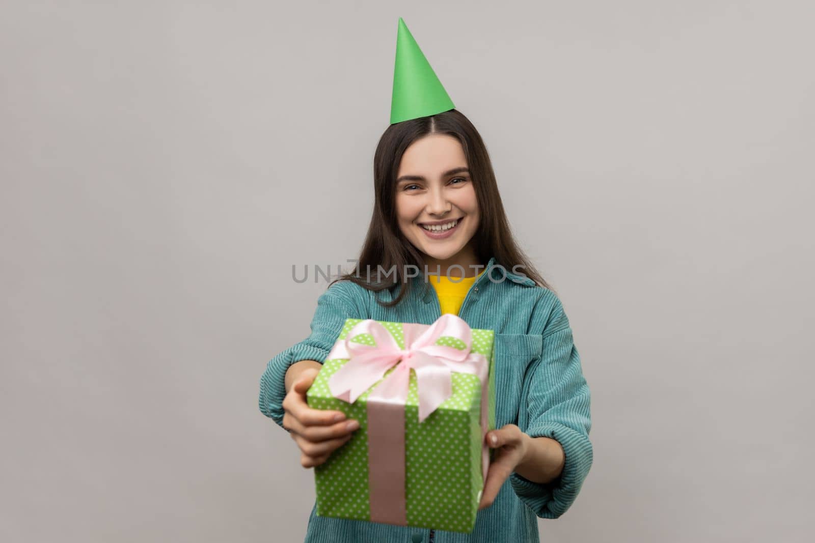 Portrait of happy woman giving green gift box to camera and smiling, sharing holiday present, congratulating on birthday, wearing casual style jacket. Indoor studio shot isolated on gray background.