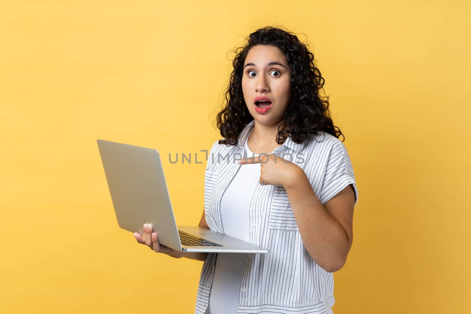 Portrait of amazed astonished woman with dark wavy hair standing with laptop in hands and pointing at display, looking with big eyes and open mouth. Indoor studio shot isolated on yellow background.