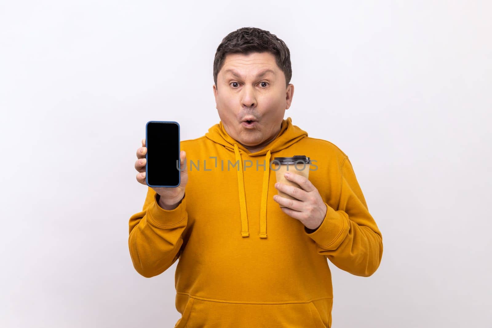 Portrait of astonished surprised man of middle age holding smart phone with blank screen and disposable cup of coffee, wearing urban style hoodie. Indoor studio shot isolated on white background.