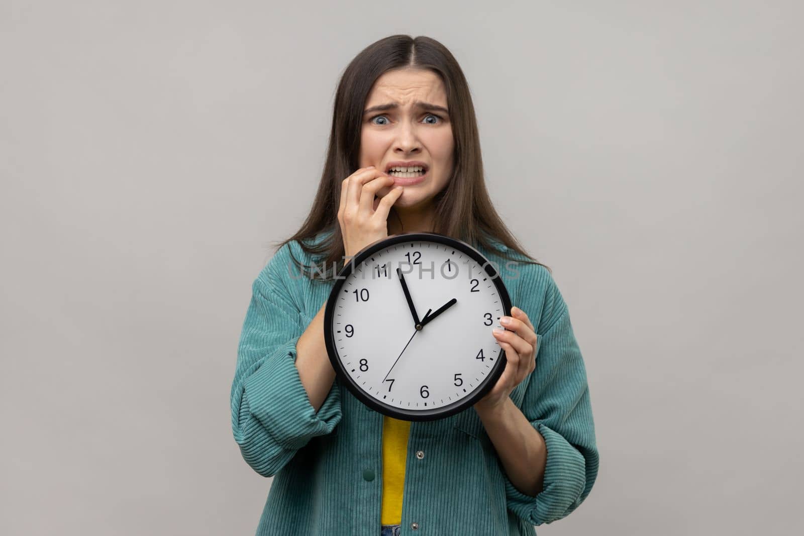 Portrait of impatient nervous woman with dark hair biting her nails and holding big wall clock, deadline, wearing casual style jacket. Indoor studio shot isolated on gray background.