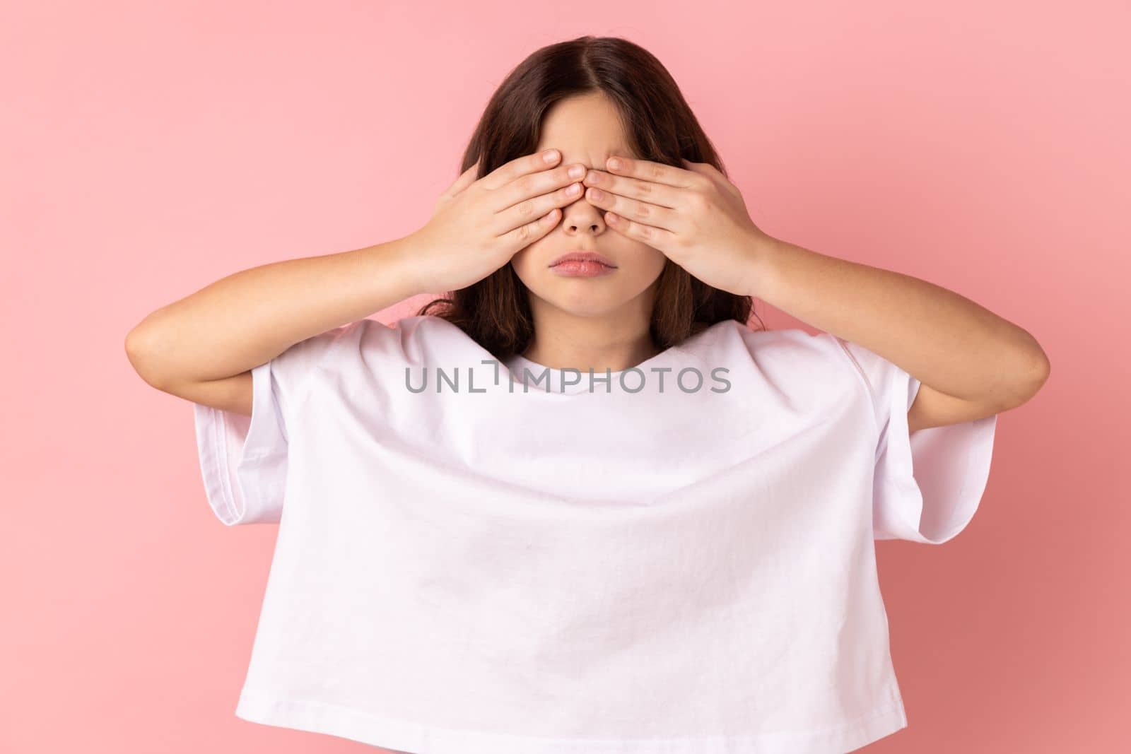 Portrait of sad upset little girl wearing white T-shirt standing covering eyes with hand, does not want see at something shameful. Indoor studio shot isolated on pink background.