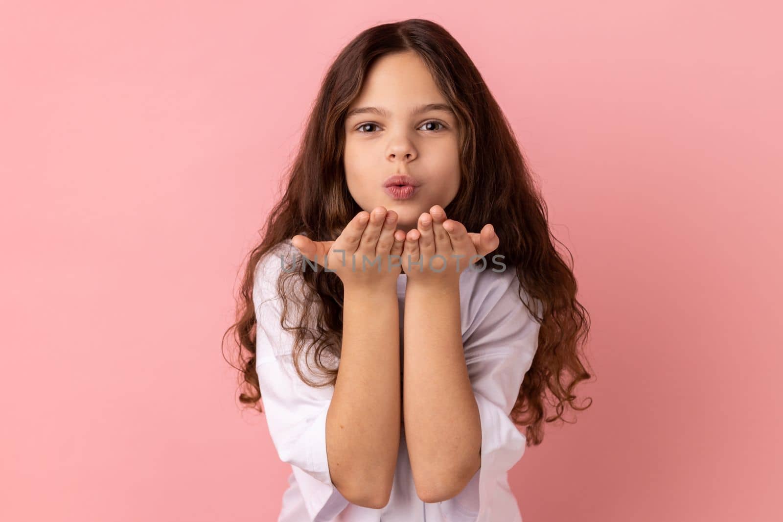 Portrait of romantic lovely little girl wearing white T-shirt sending air kisses to her friend, looking at camera, falling in love. Indoor studio shot isolated on pink background.