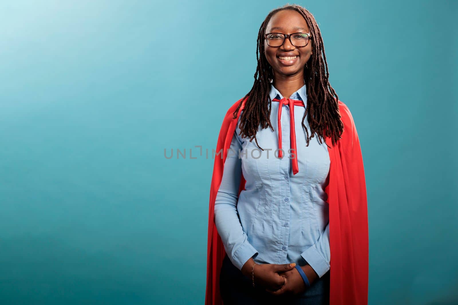 Positive smiling heartily mighty and brave justice defender standing on blue background while looking at camera. Young adult confident and strong superhero woman wearing hero cloak. Studio shot