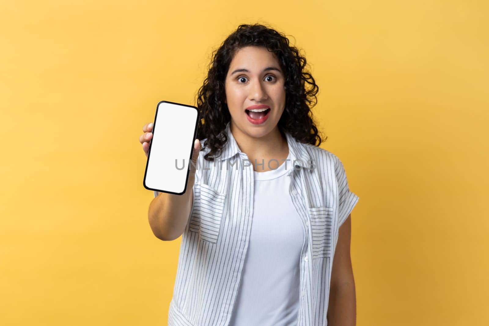 Portrait of excited amazed woman with dark wavy hair holding phone with empty display, advertisement area, copy space for promotional text. Indoor studio shot isolated on yellow background.