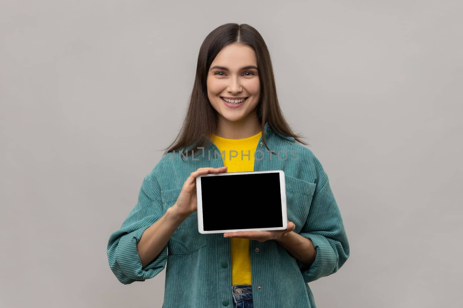 Young smiling woman holding digital tablet in hands, showing blank device screen to camera, expressing happiness, wearing casual style jacket. Indoor studio shot isolated on gray background.