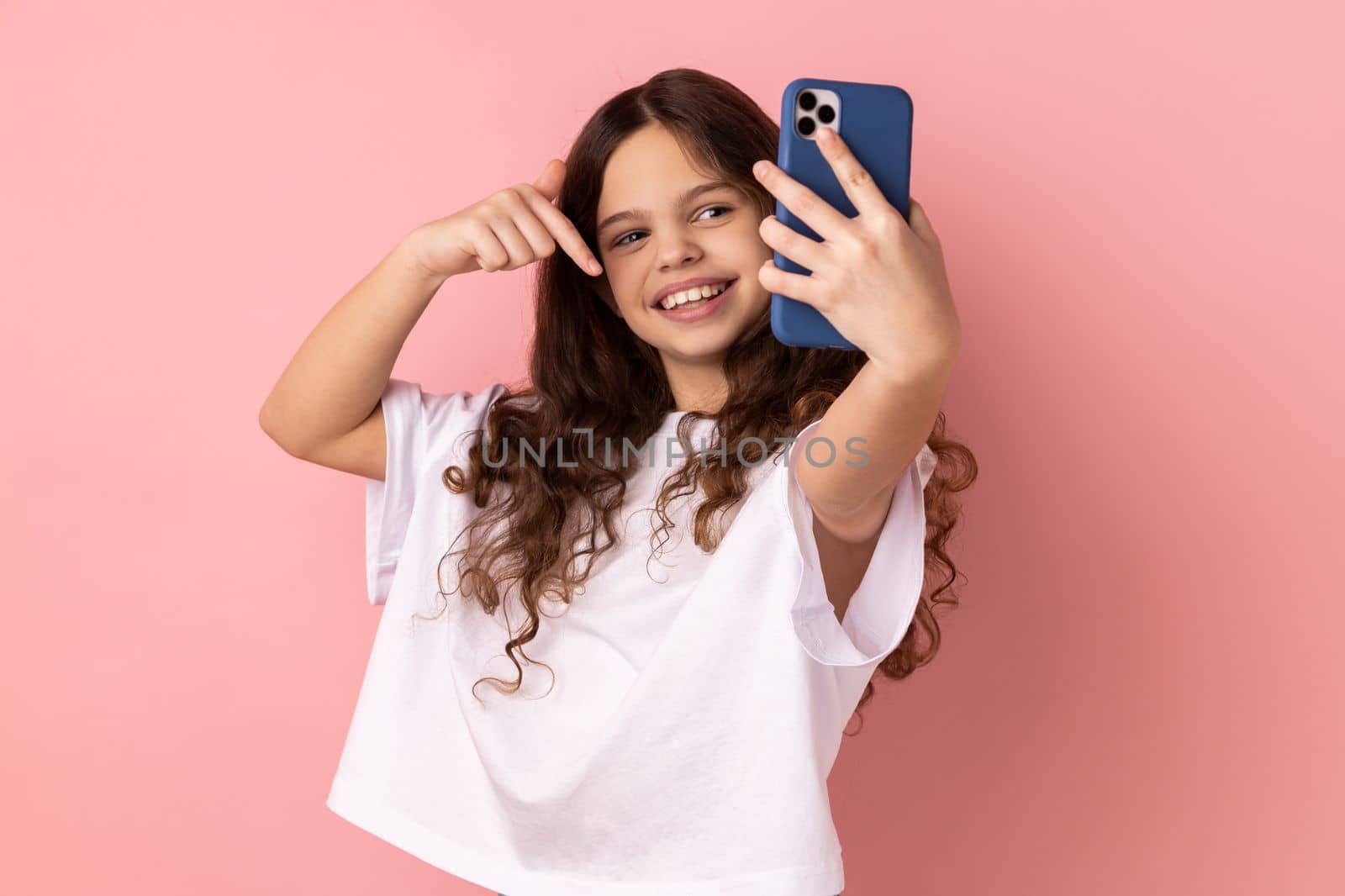 Little girl wearing white T-shirt pointing down while communicating by video call or streaming, gesturing to subscribe, taking selfie on smartphone. Indoor studio shot isolated on pink background.