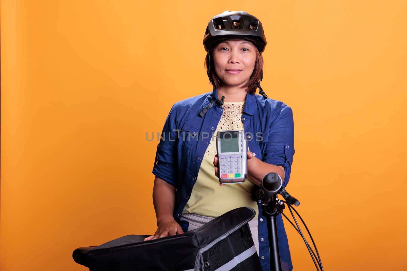 Portrait of deliverywoman with blue uniform and helmet delivering fast food order giving POS terminal to customer. Pizzeria employee using bike as transportation while carrying takeout food backpack